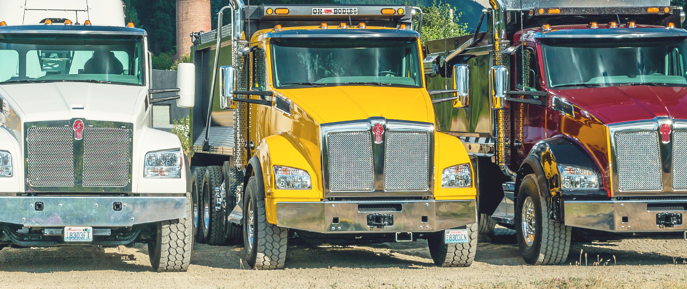 A row of Kenworth T880 trucks parked on dirt with factory in background